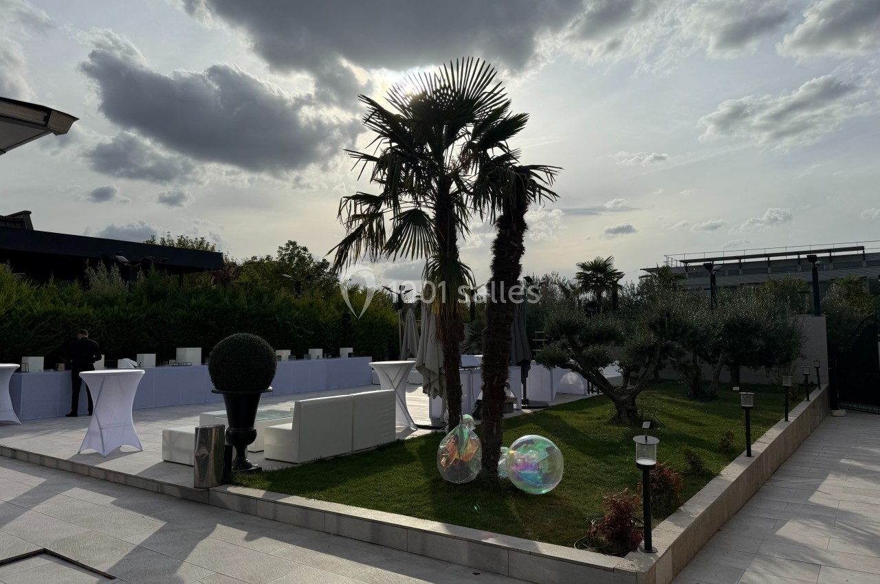 Jardin aménagé avec palmiers, tables hautes blanches et ballons transparents sous un ciel partiellement nuageux.