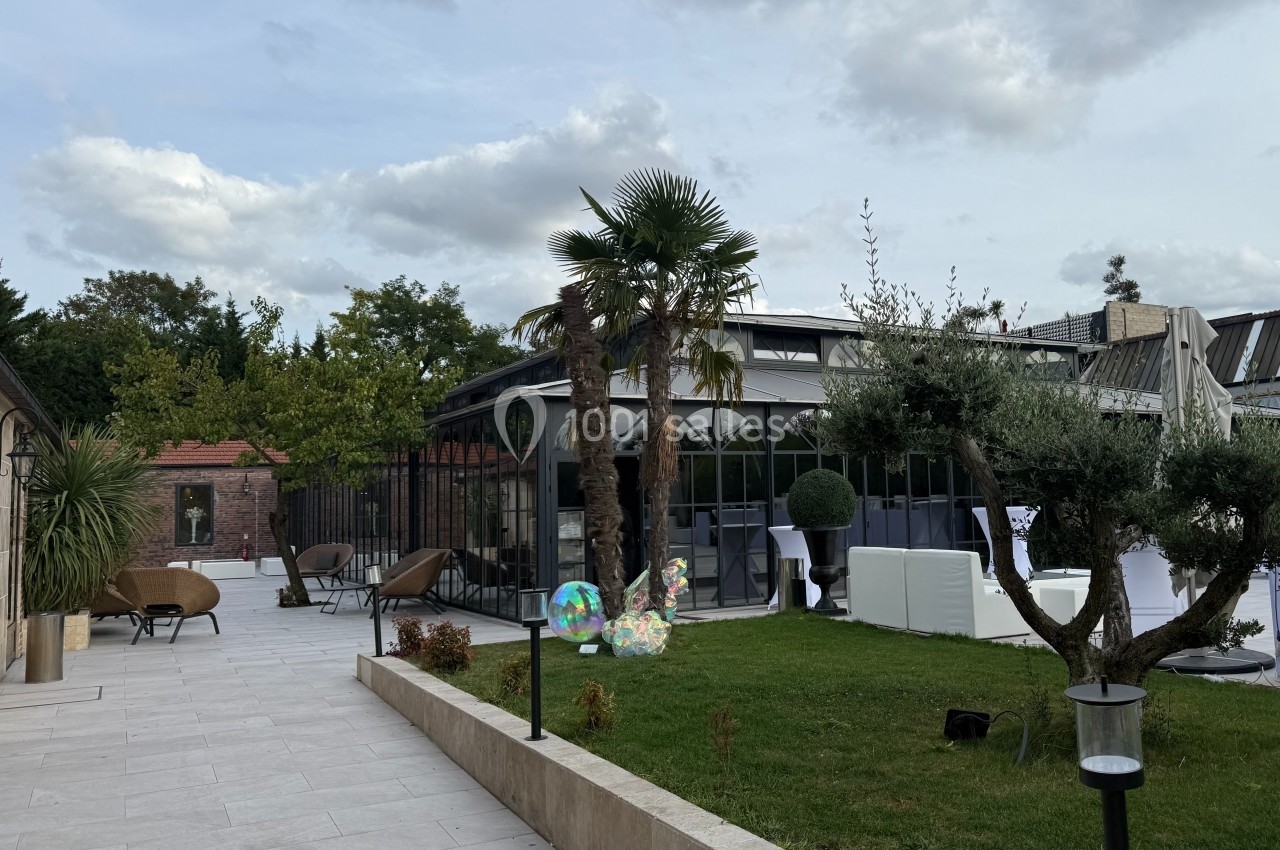 Terrasse avec dallage clair, jardin arboré, mobilier extérieur et bâtiment vitré sous un ciel partiellement nuageux.