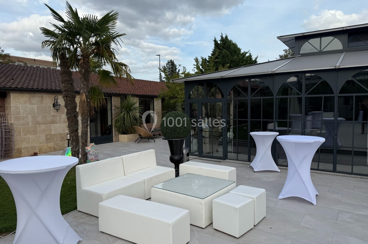 Mobilier de salon blanc avec table basse en verre sur une terrasse extérieure, entourée de végétation et d'une véranda.