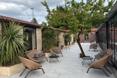 Terrasse extérieure avec tables, chaises, parasol, plantes et vue sur un bâtiment vitré sous un ciel dégagé.