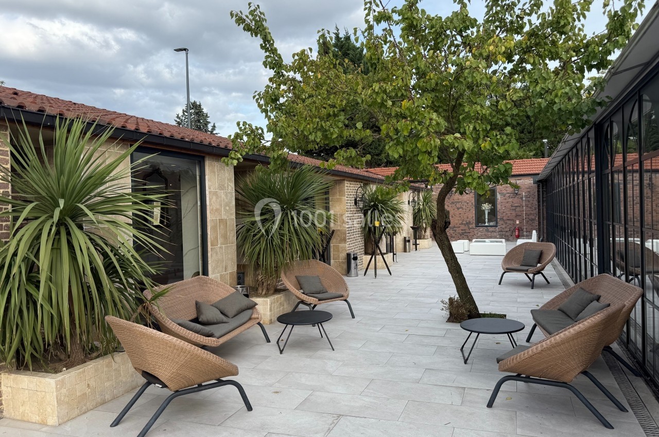 Terrasse aménagée avec fauteuils en rotin, tables basses, plantes et un arbre, entourée de bâtiments en briques et verre.