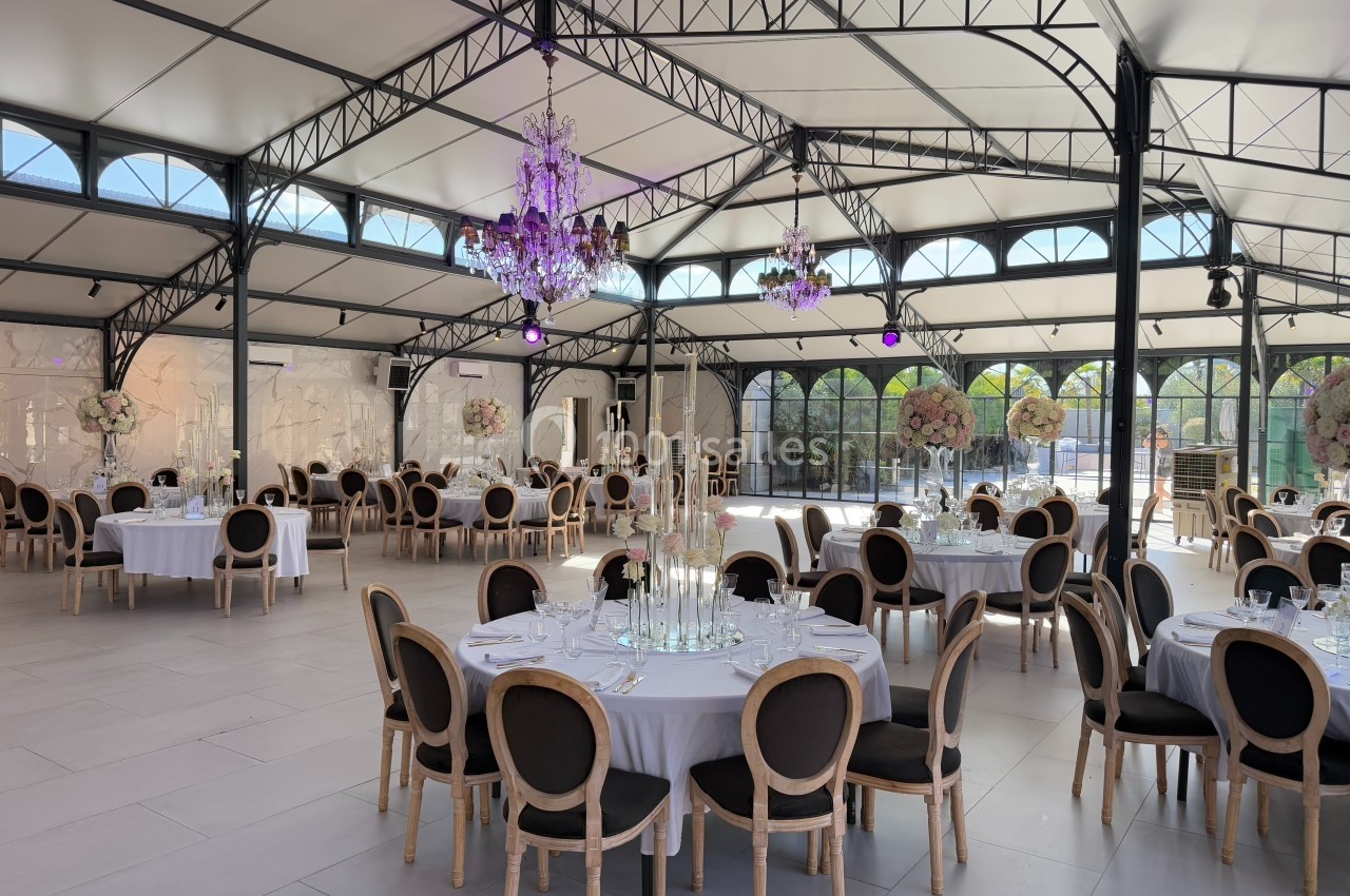 Salle de réception lumineuse avec tables rondes dressées, chaises élégantes et lustres suspendus.
