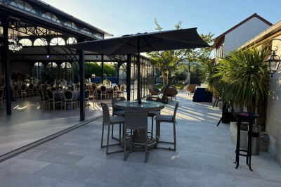 Terrasse extérieure avec tables, chaises, parasol, plantes et vue sur un bâtiment vitré sous un ciel dégagé.