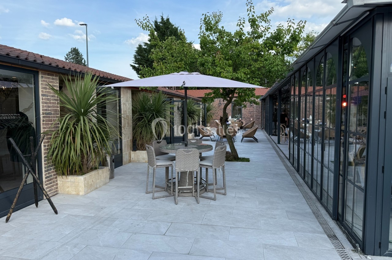 Terrasse extérieure avec tables, chaises, parasol, plantes et vue sur un bâtiment vitré sous un ciel dégagé.