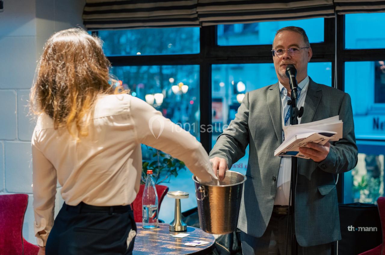 Un homme tenant un micro supervise une femme tirant un papier d'un seau dans une salle éclairée.