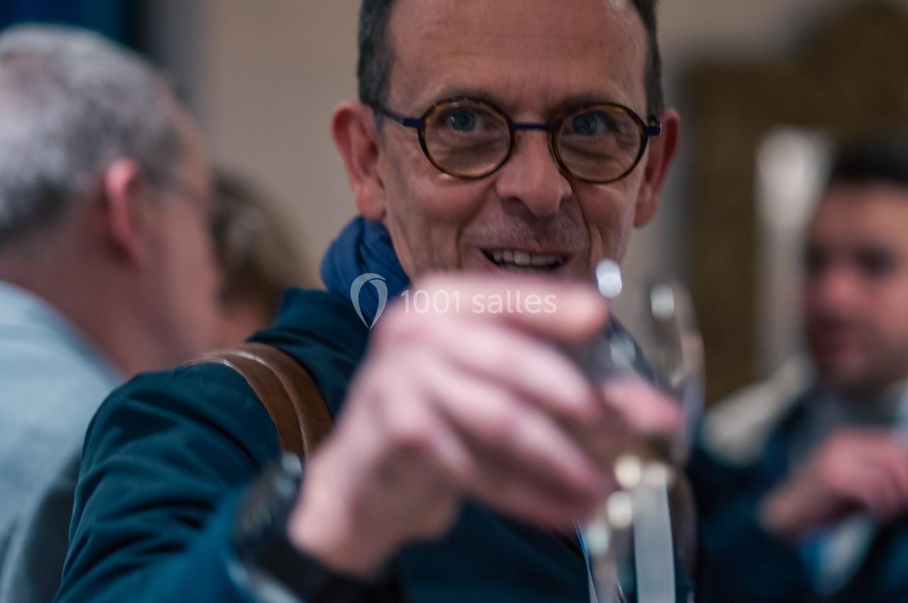 Homme portant des lunettes, tenant un verre à la main dans un cadre convivial avec des personnes en arrière-plan.