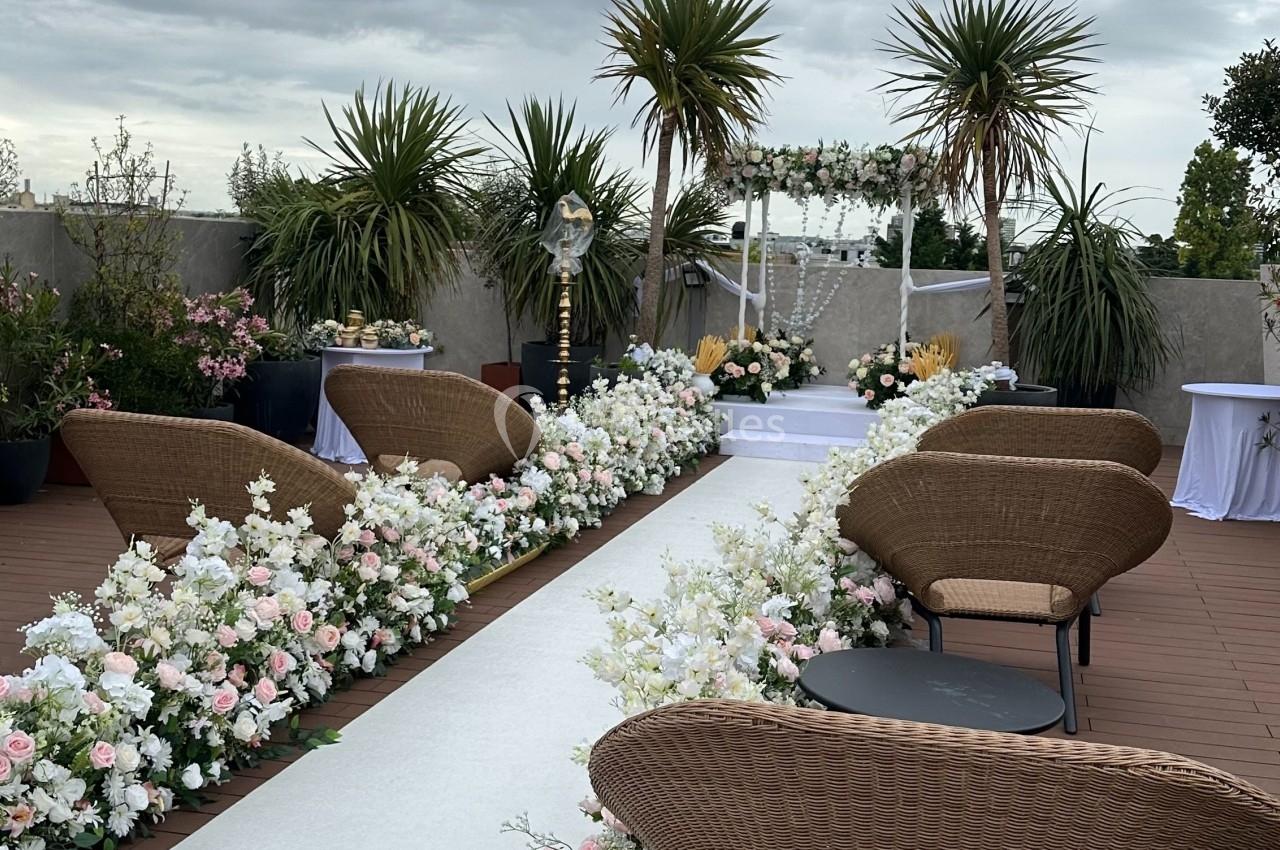 Terrasse aménagée avec des chaises en rotin, allée blanche bordée de fleurs et ciel nuageux en arrière-plan.