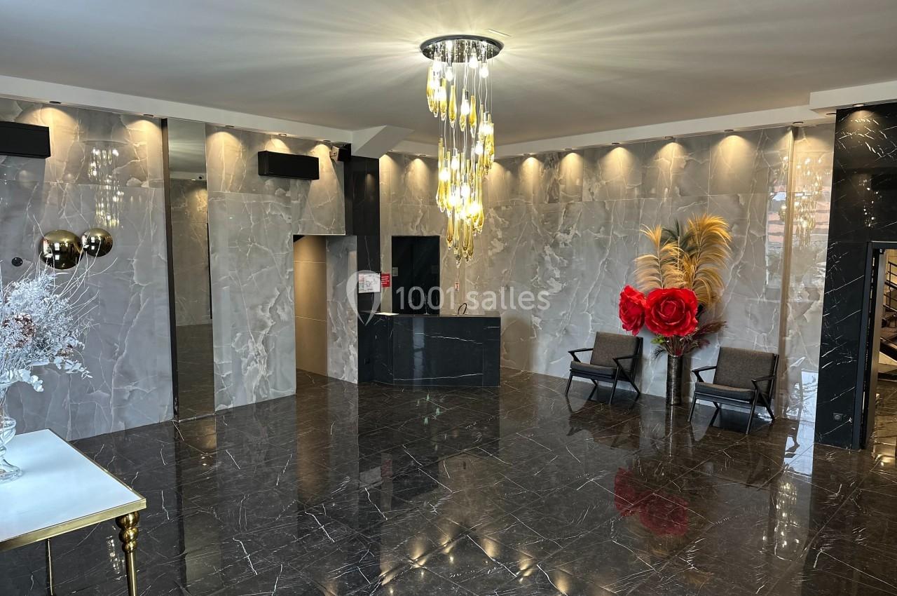 Hall moderne avec sol en marbre noir, murs clairs, lustre design, fauteuils, et décoration florale rouge et dorée.