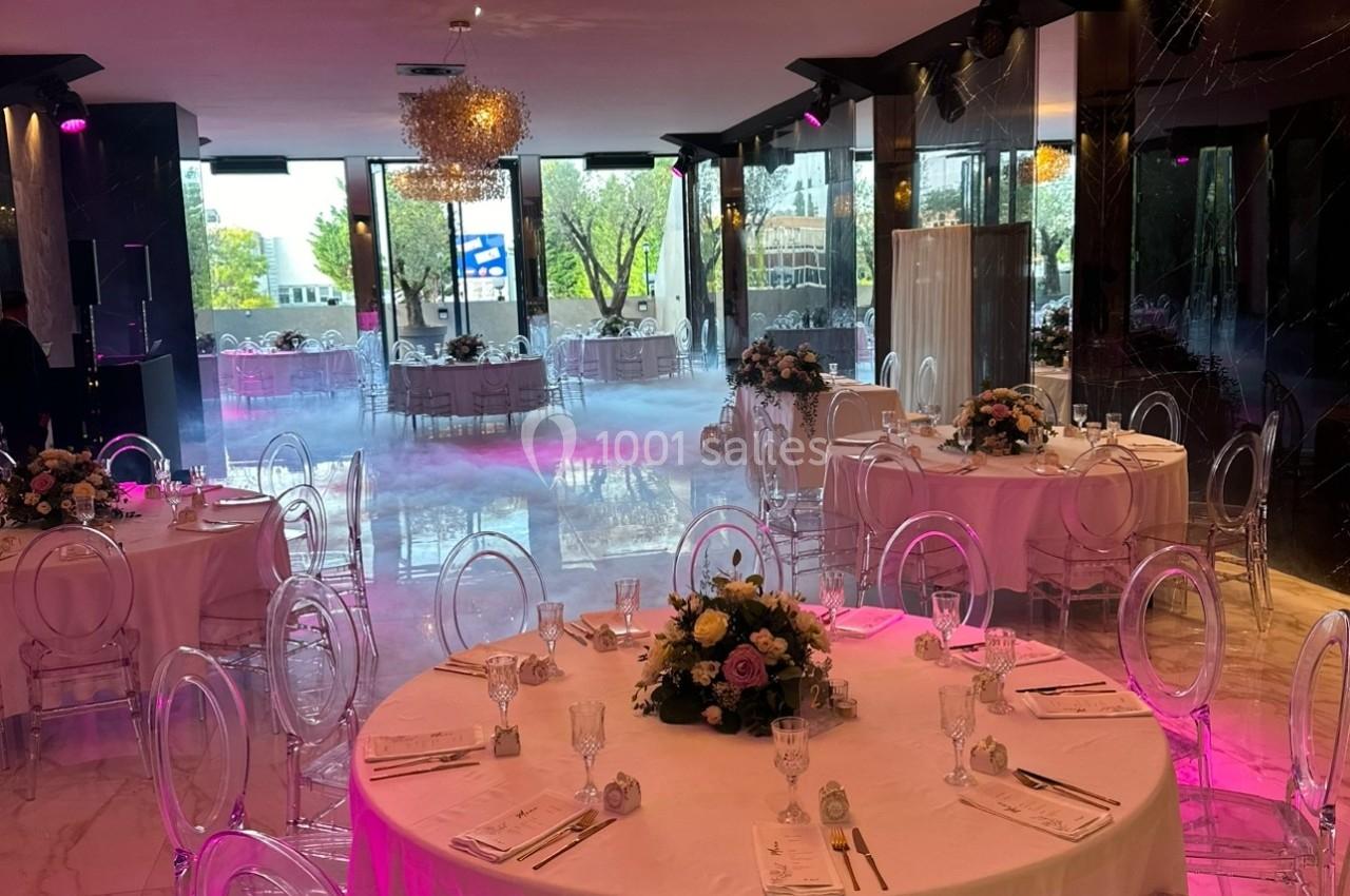 Salle de réception élégante avec tables rondes dressées, chaises transparentes et lustres dorés, éclairage tamisé.