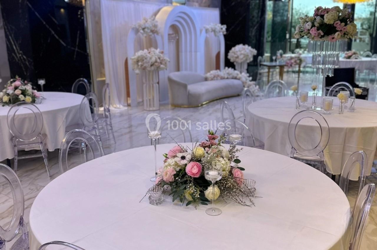 Salle de réception élégante avec tables rondes décorées de bouquets floraux et une arche ornée en arrière-plan.