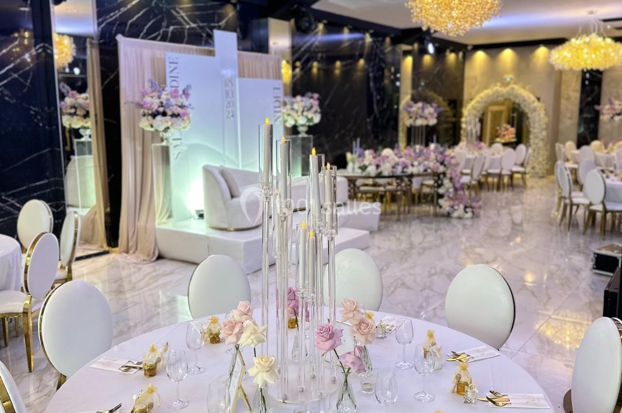 Salle de réception élégante avec tables rondes décorées de bougies, fleurs pastel et couverts dorés.