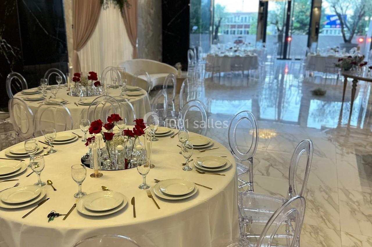 Salle de réception élégante avec tables rondes dressées, chaises transparentes et décorations florales rouges.