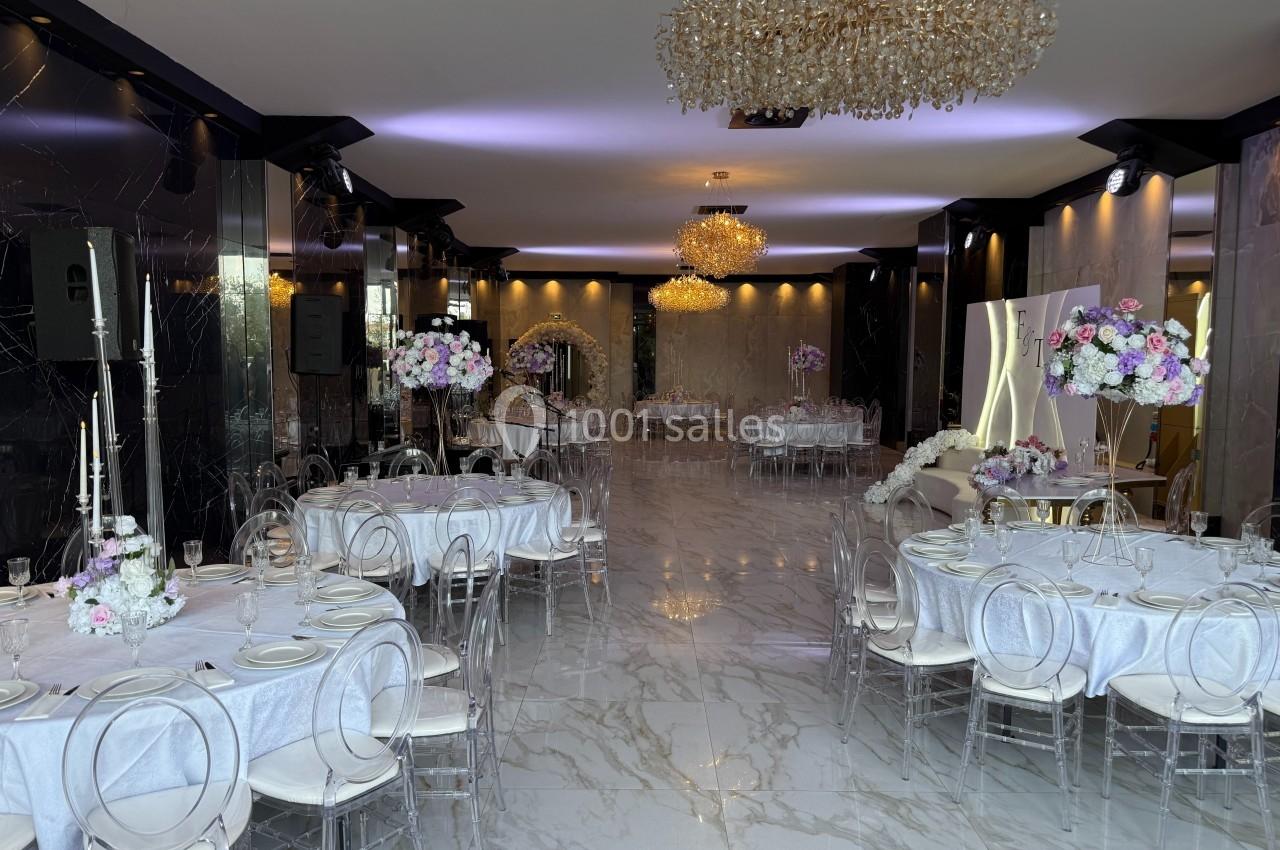 Salle de réception élégante avec tables dressées, chaises transparentes et lustres en cristal.