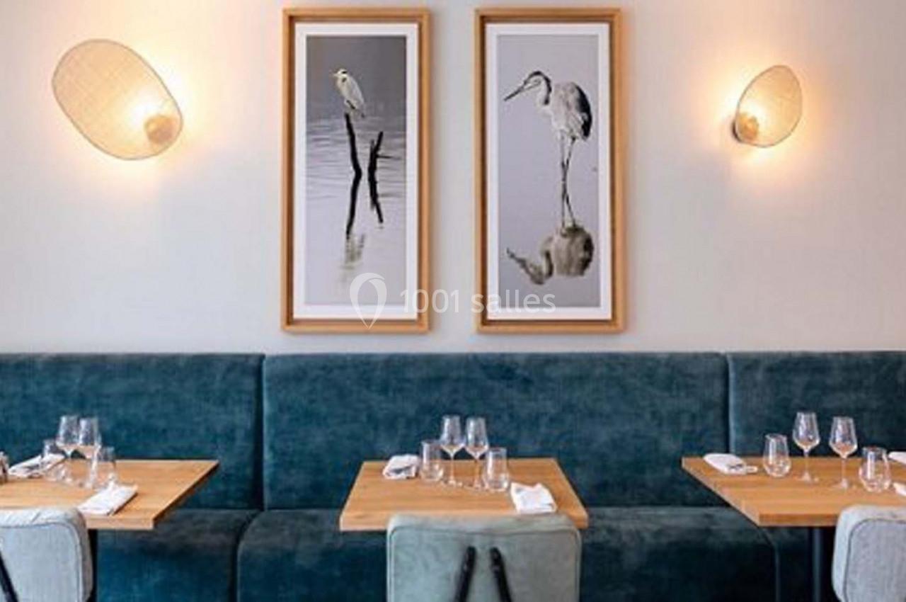Tables en bois dressées avec verres et serviettes devant une banquette bleue, décorée de cadres et appliques murales.