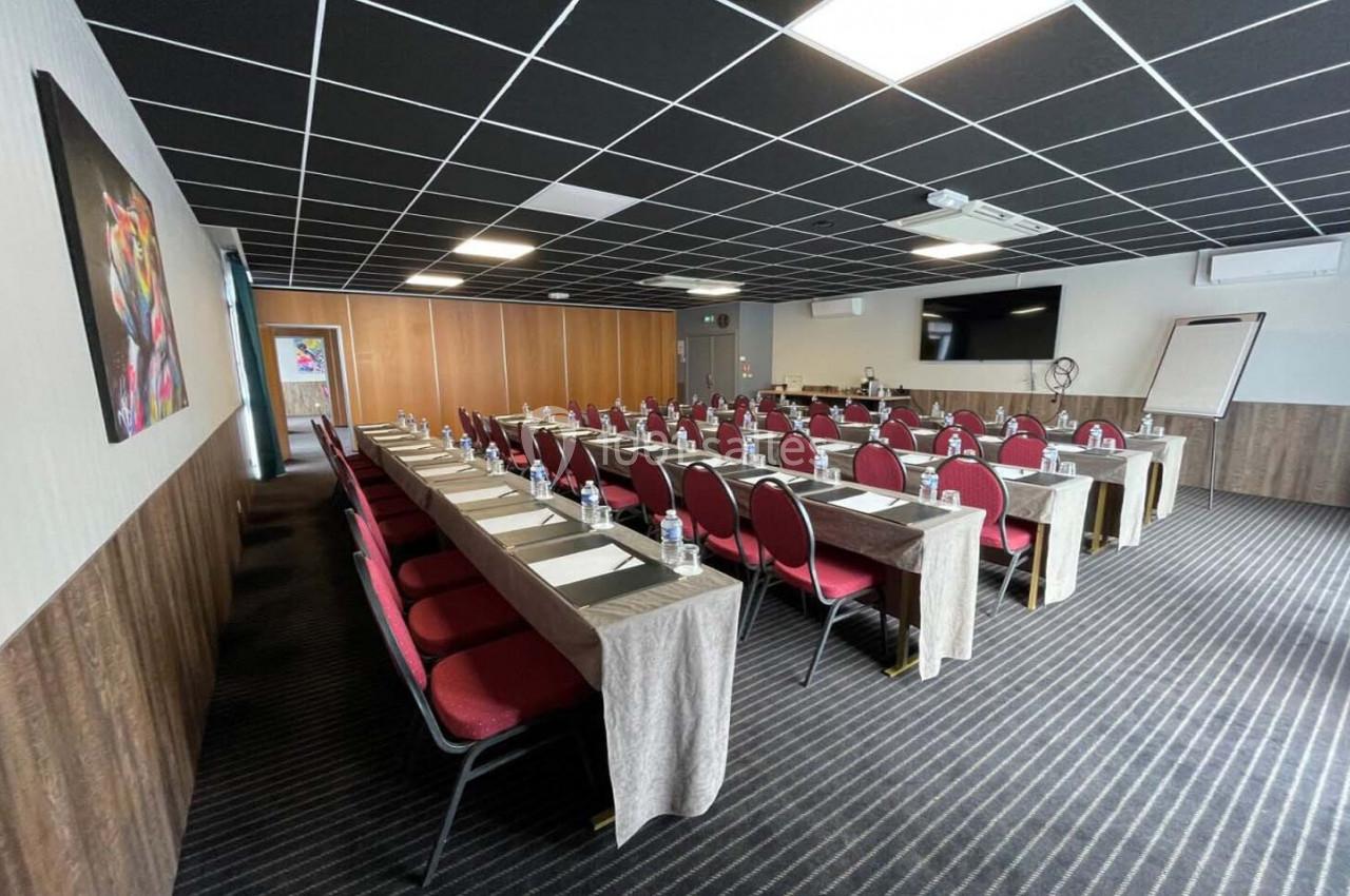 Salle de réunion avec tables disposées en rangées, chaises rouges, écran mural, paperboard et éclairage au plafond.