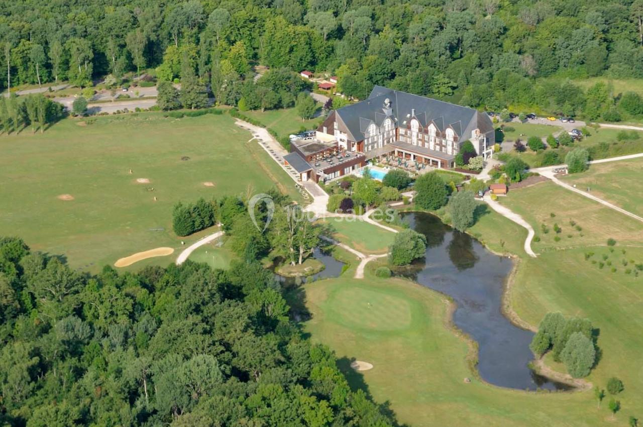 Vue aérienne d'un domaine avec un grand bâtiment, des espaces verts, un étang et des chemins entourés de forêt.