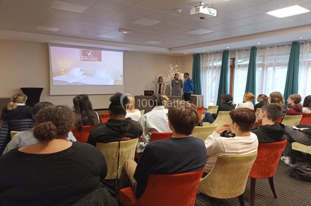 Un groupe de personnes assises dans une salle de conférence, écoutant une présentation projetée sur un écran.