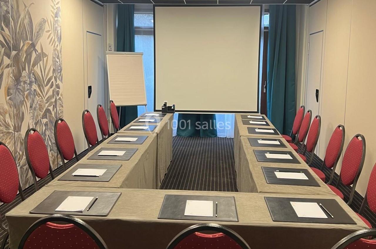 Salle de réunion avec disposition en U, chaises rouges, tables couvertes de nappes beiges, écran de projection et paperboard.