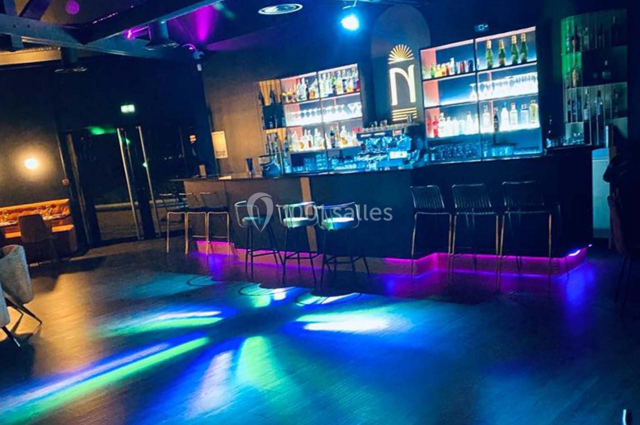 Bar moderne avec comptoir éclairé, tabourets alignés et jeux de lumières colorées sur le sol sombre.