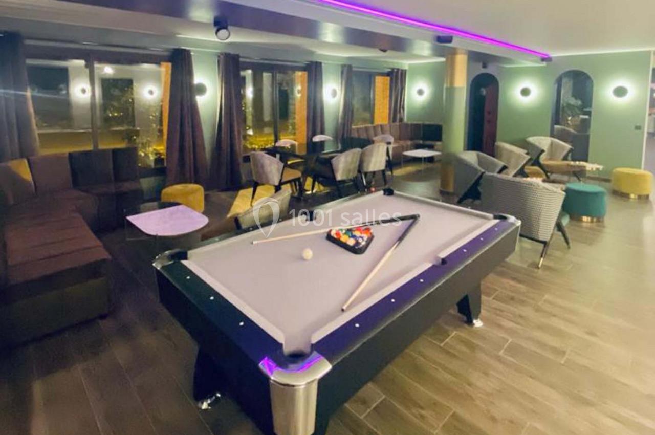 Salle moderne avec table de billard éclairée, entourée de fauteuils, tables basses et grandes fenêtres donnant sur l…