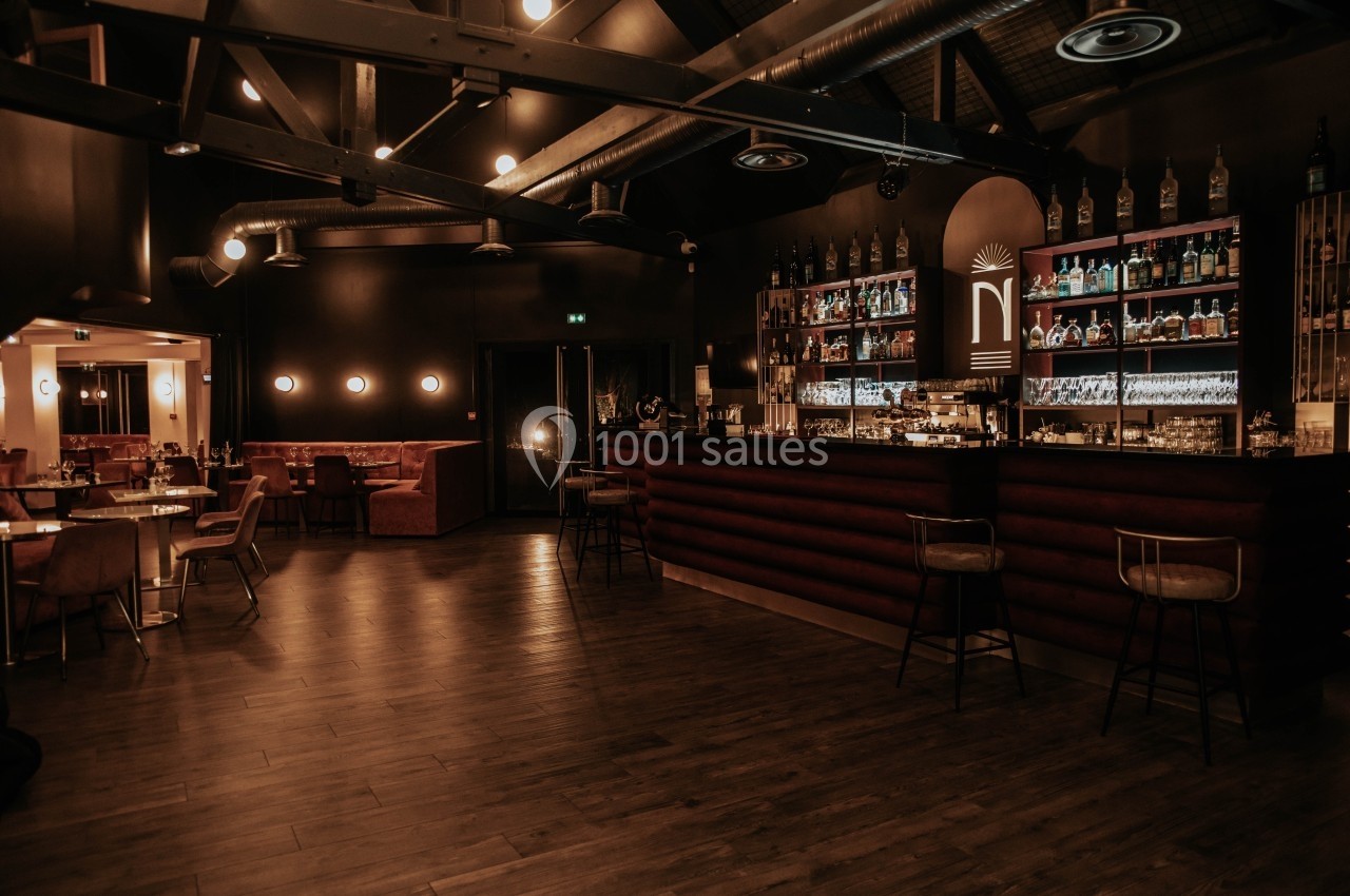 Intérieur d'un bar élégant avec comptoir éclairé, étagères de bouteilles et tables en bois dans une ambiance tamisée.