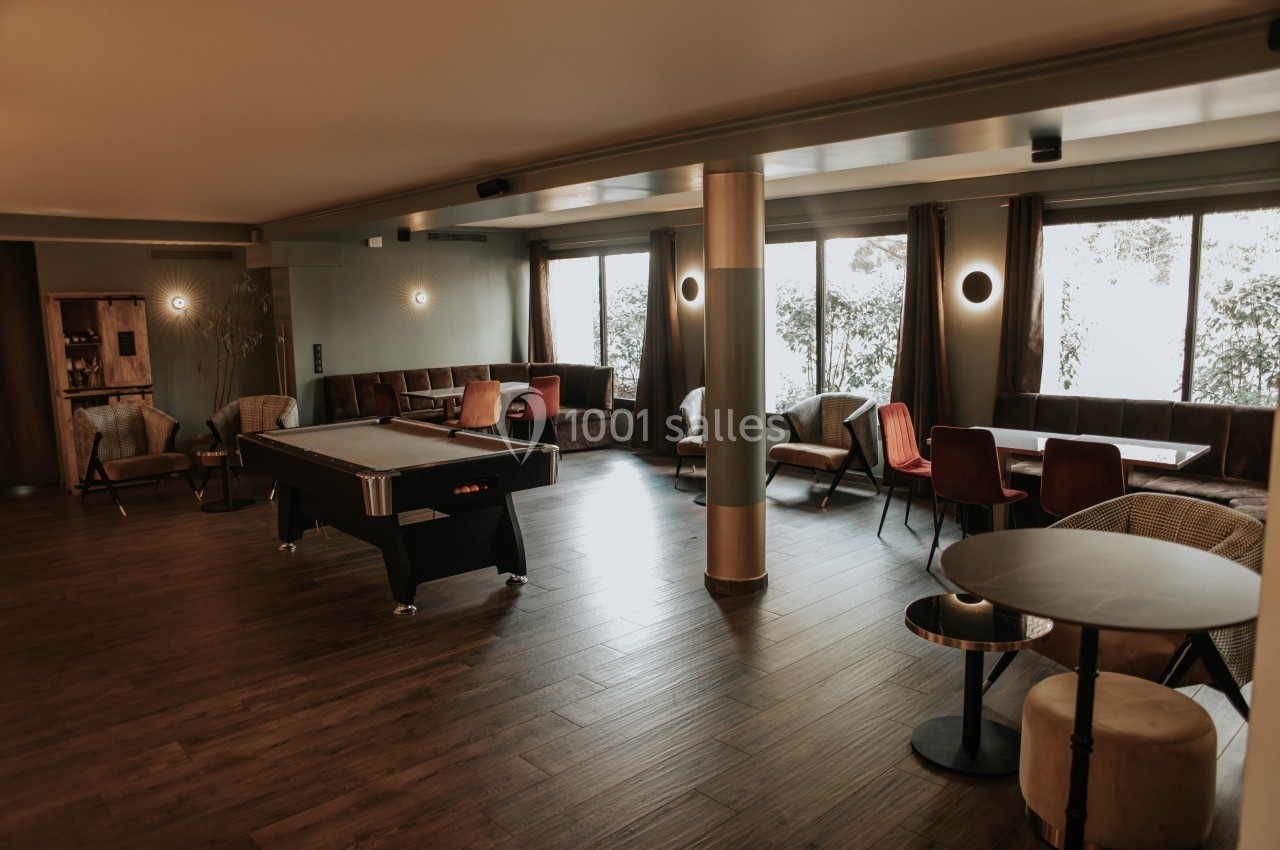 Salle lumineuse avec parquet, table de billard, fauteuils et tables entourées de grandes fenêtres donnant sur un jardin.