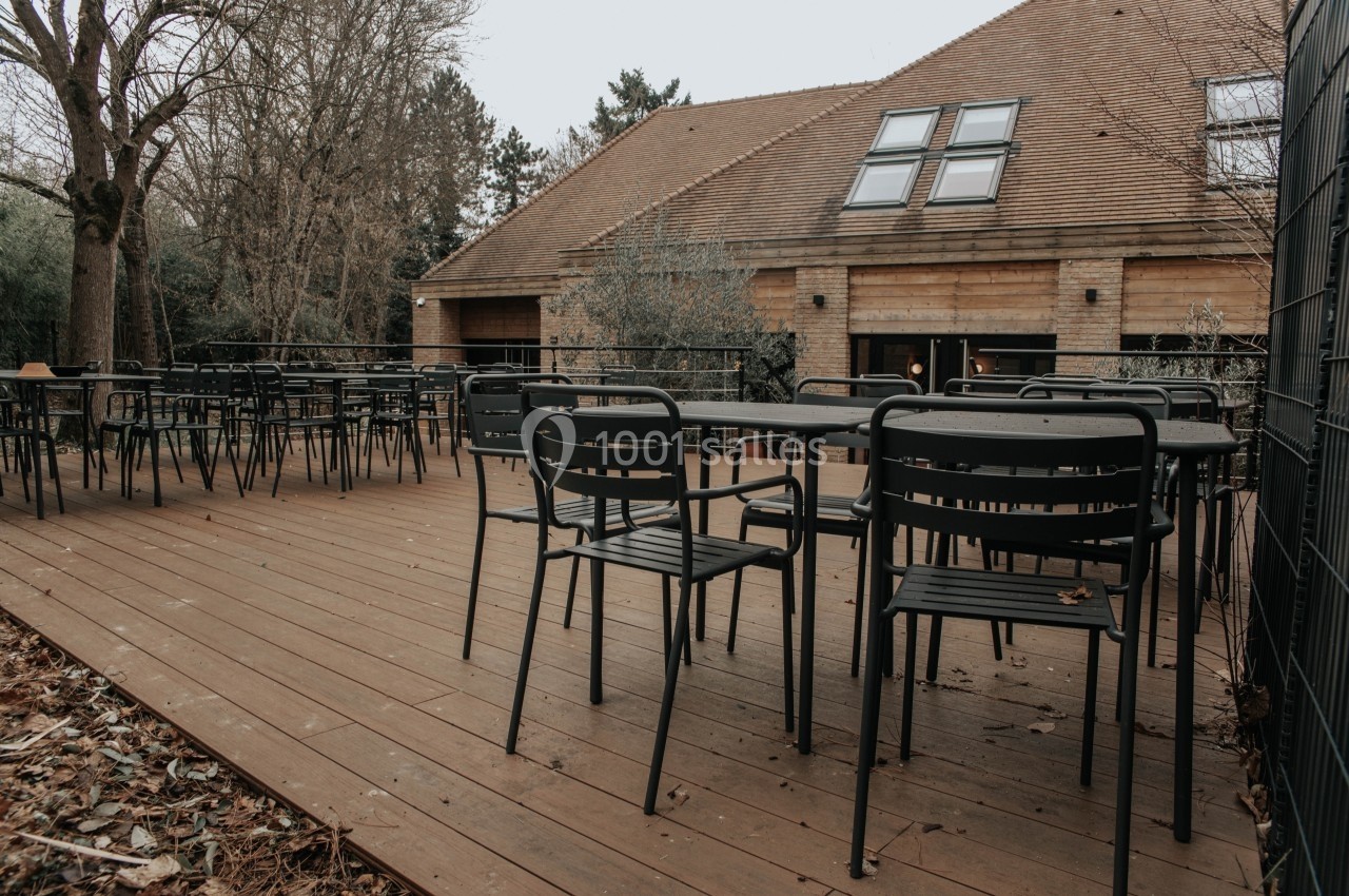Terrasse en bois avec tables et chaises noires, entourée d'arbres et donnant sur un bâtiment en pierre et bois.