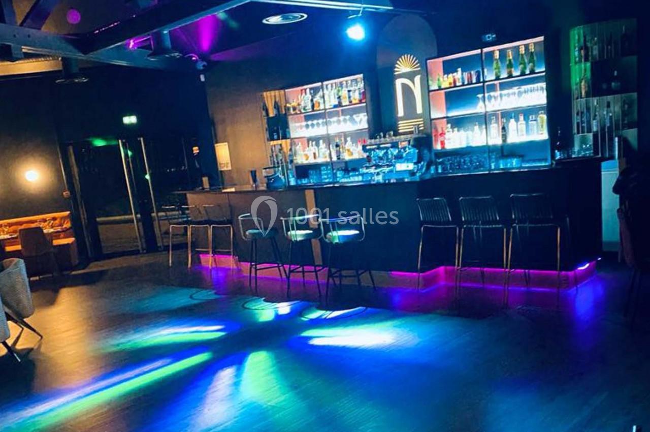 Bar moderne avec comptoir éclairé, tabourets hauts et jeux de lumières colorées sur le sol sombre.