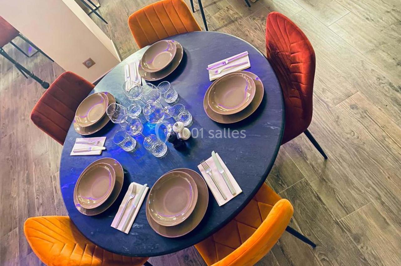 Table ronde dressée avec six assiettes, couverts et verres, entourée de chaises orange et jaunes sur un sol en bois.
