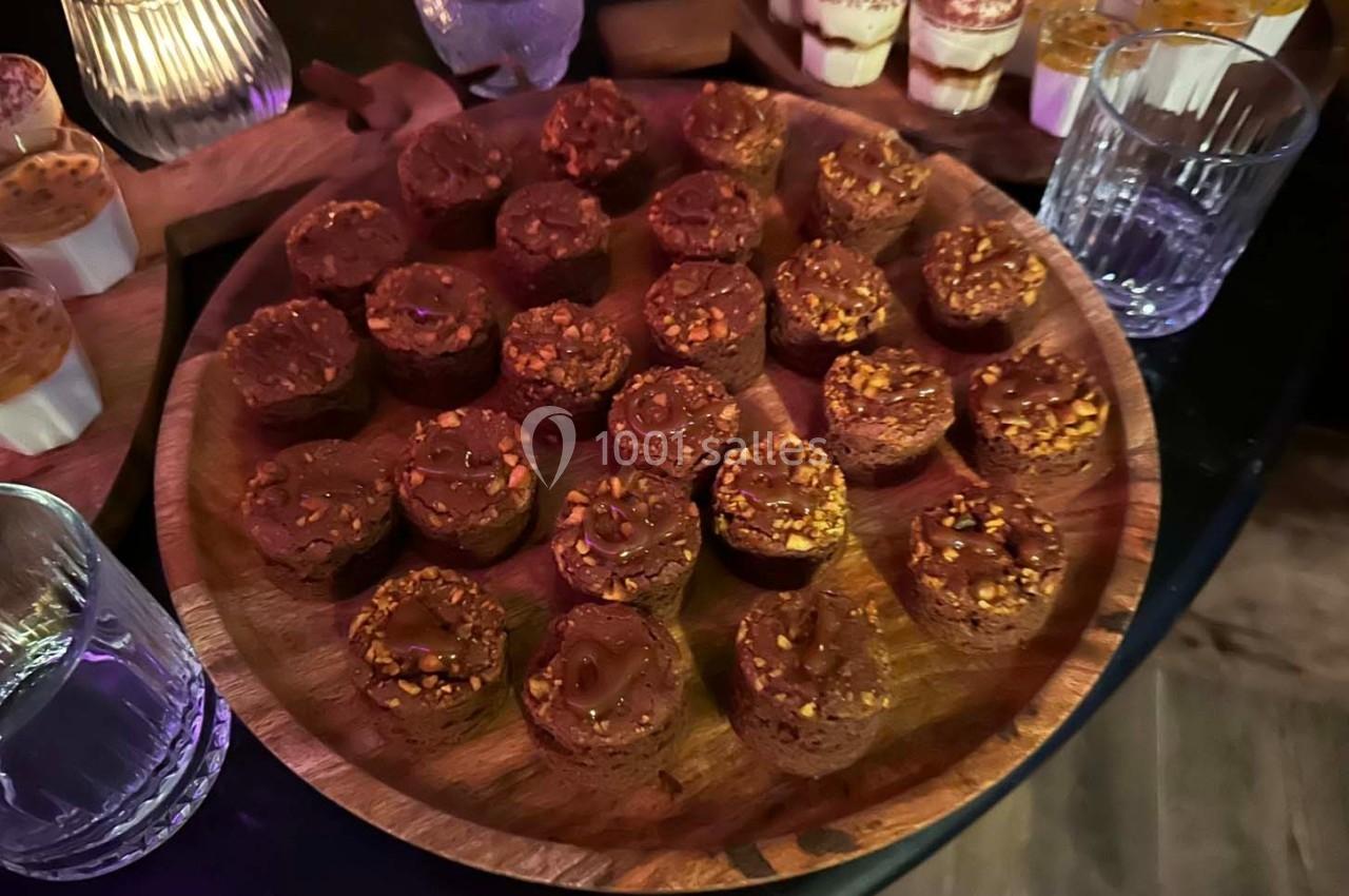 Plateau de petits desserts chocolatés garnis de noisettes, entouré de verrines et de verres sur une table sombre.
