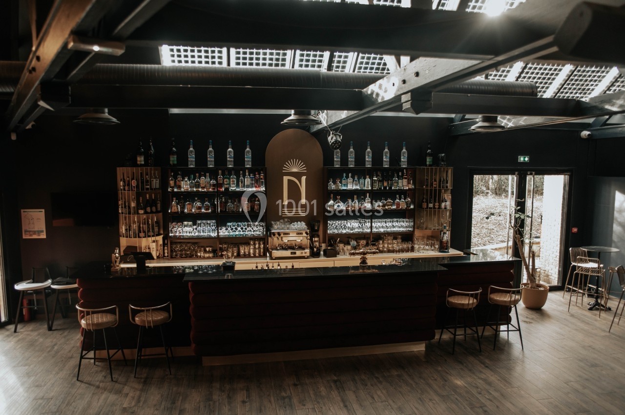 Bar moderne avec comptoir central, étagères garnies de bouteilles et lumière naturelle entrant par le toit vitré.