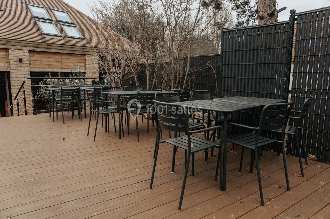 Terrasse en bois avec plusieurs tables et chaises noires, entourée de clôtures et de végétation hivernale.