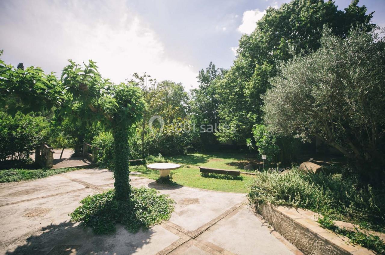 Jardin verdoyant avec arbres, arbustes, table en pierre et allées pavées sous un ciel partiellement nuageux.