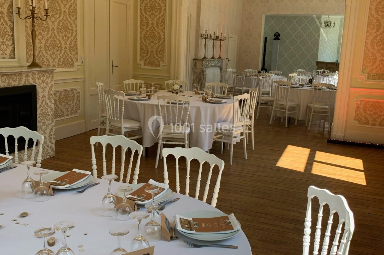 Salle de réception élégante avec tables rondes dressées, chaises blanches et lumière naturelle entrant par les fenêtres.