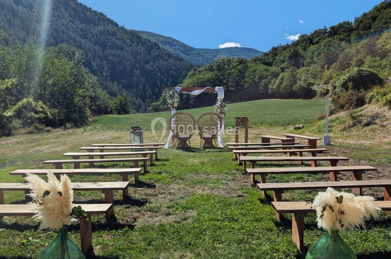Chaises et bancs disposés en extérieur pour une cérémonie, avec une arche décorée au fond, entourés de montagnes verdoyantes.