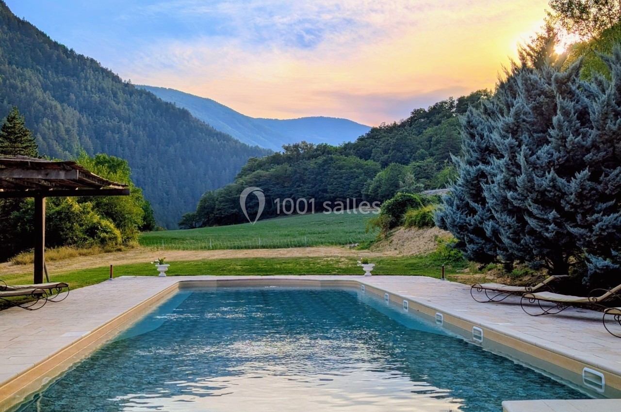 Piscine extérieure entourée de chaises longues, avec vue sur des collines boisées et un ciel coloré au coucher du soleil.