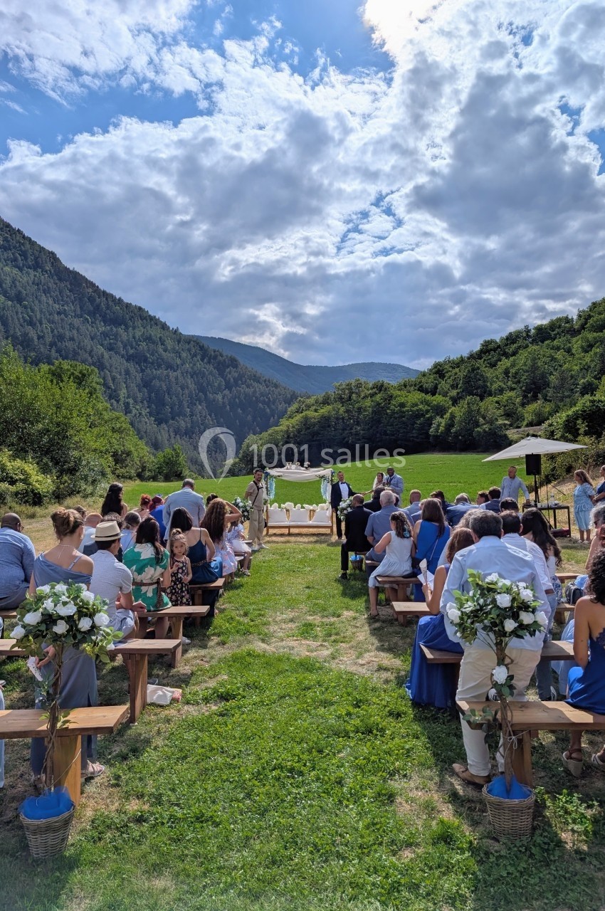 Cérémonie de mariage en plein air dans une vallée montagneuse, avec invités assis sur des bancs en bois.