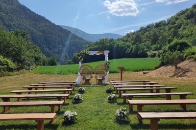 Un homme en costume parle devant une arche décorée de fleurs séchées, dans un cadre naturel avec des montagnes et des arbres.