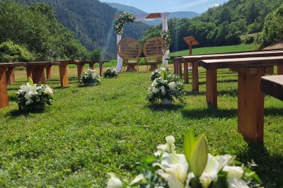 Un homme en costume parle devant une arche décorée de fleurs séchées, dans un cadre naturel avec des montagnes et des arbres.