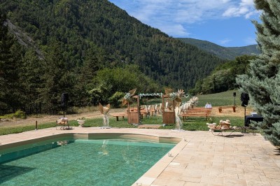 Un homme en costume parle devant une arche décorée de fleurs séchées, dans un cadre naturel avec des montagnes et des arbres.