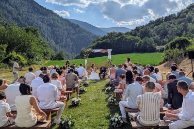 Un homme en costume parle devant une arche décorée de fleurs séchées, dans un cadre naturel avec des montagnes et des arbres.