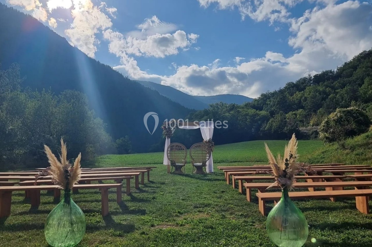 Cérémonie en plein air avec arche décorée, bancs en bois et montagnes en arrière-plan sous un ciel ensoleillé.