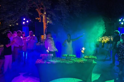 Un homme en costume parle devant une arche décorée de fleurs séchées, dans un cadre naturel avec des montagnes et des arbres.
