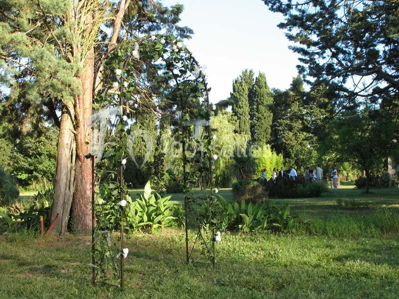 Arche décorée de feuillage et de fleurs blanches dans un jardin verdoyant avec des arbres et des promeneurs au loin.