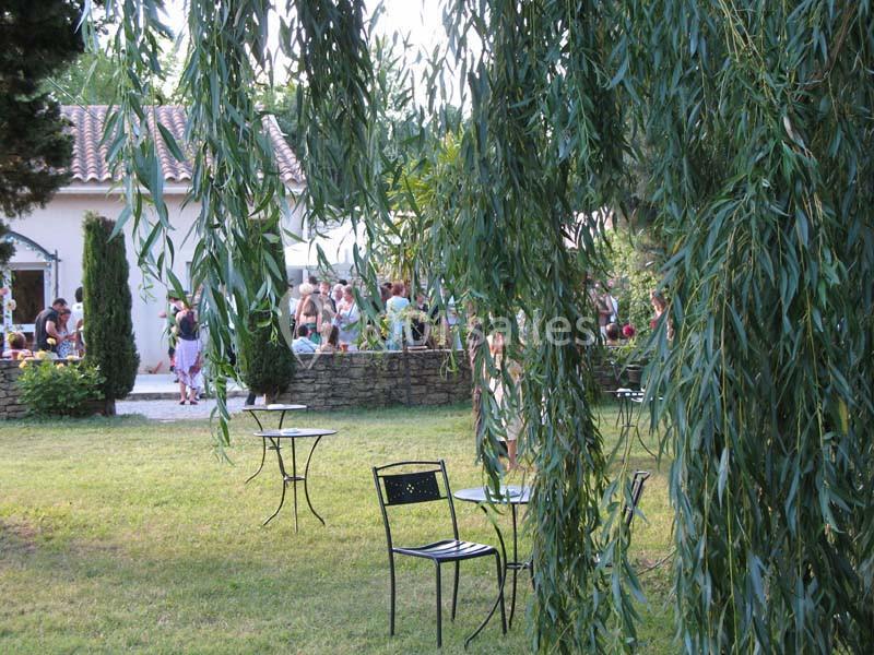 Des tables et chaises en métal noir dans un jardin verdoyant, avec des personnes réunies près d'une maison en arrière-plan.
