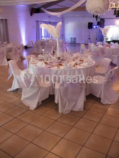 Salle de réception décorée avec des tables rondes, nappes blanches, chaises couvertes et centre de table avec plumes…