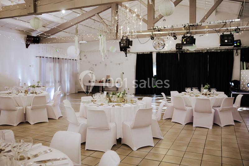 Salle de réception décorée avec des tables rondes dressées, des chaises blanches et des guirlandes lumineuses suspendues.