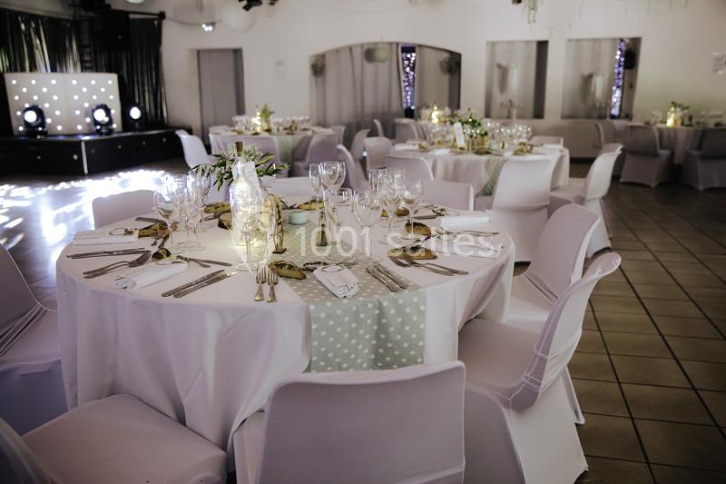 Salle de réception décorée avec des tables rondes dressées, nappes blanches, centres de table floraux et éclairage tamisé.