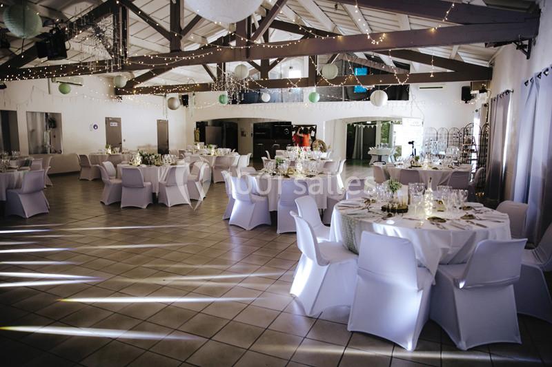 Salle de réception décorée avec des tables rondes dressées, des chaises blanches et des guirlandes lumineuses suspendues.