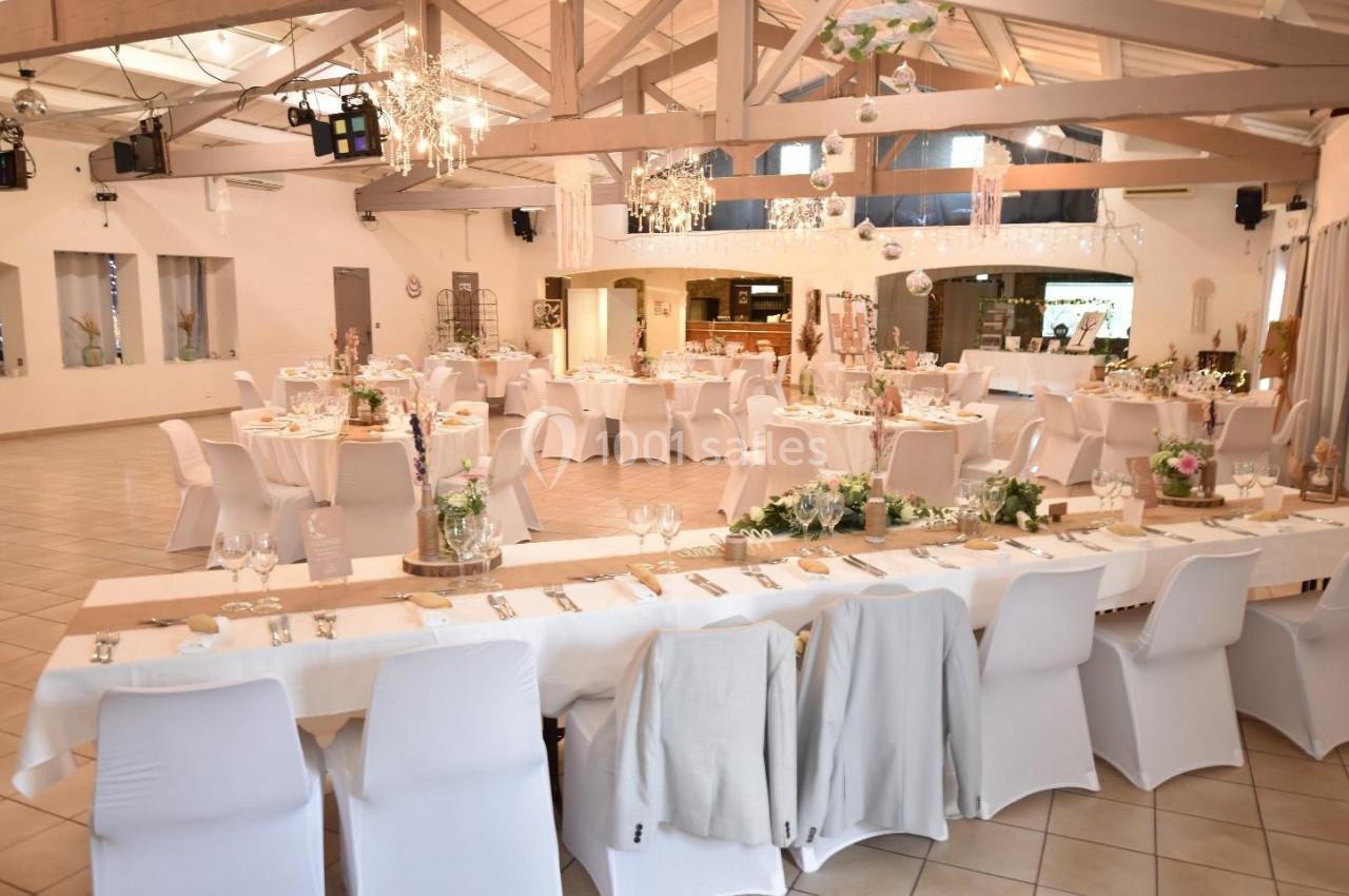 Salle de réception décorée pour un événement, avec tables dressées, chaises blanches et éclairage suspendu.